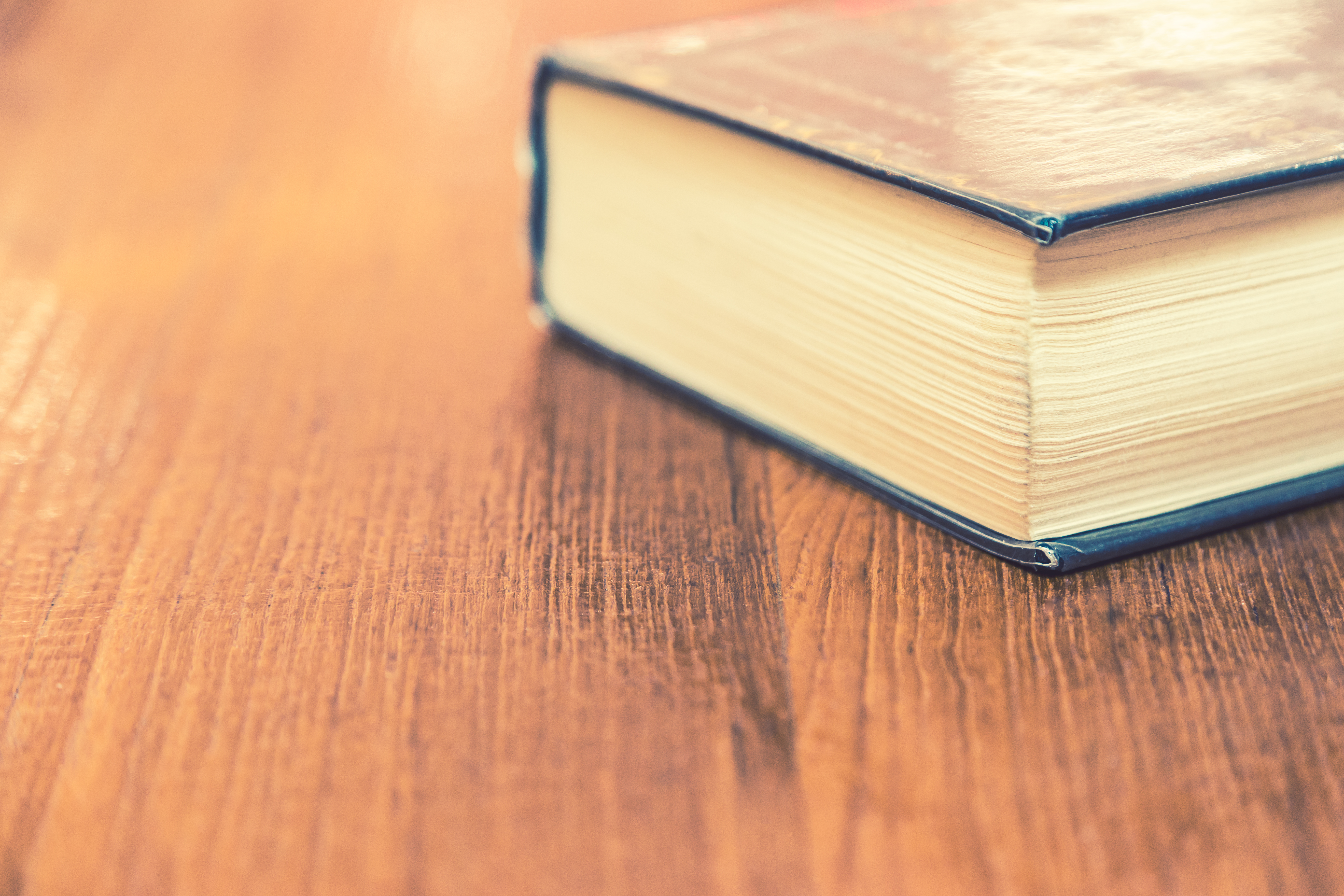 A long hardcover book lies closed on a wooden surface, its pages and spine hinting at the depth within.